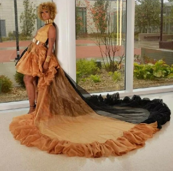 Golden-tan ruffled mini dress with a very long sheer tulle train trimmed in black, gold choker, wide gold belt and cuffs, posed by floor-to-ceiling windows