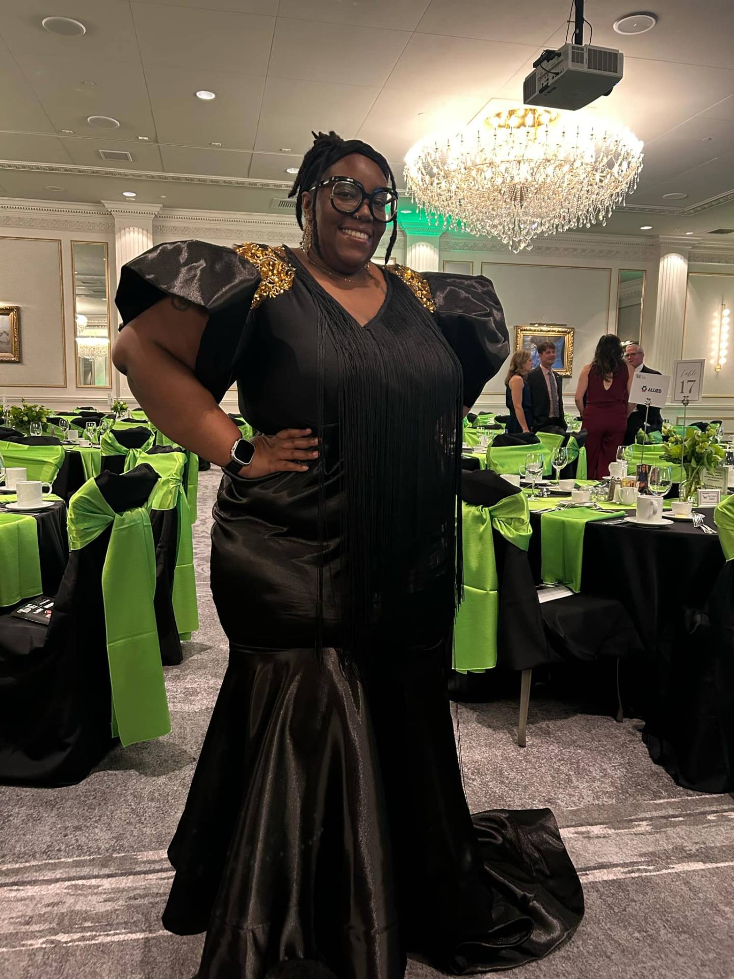 Black satin floor-length gown with puff sleeves, gold beaded shoulder detail and long black fringe draping the bodice, in a chandelier-lit ballroom