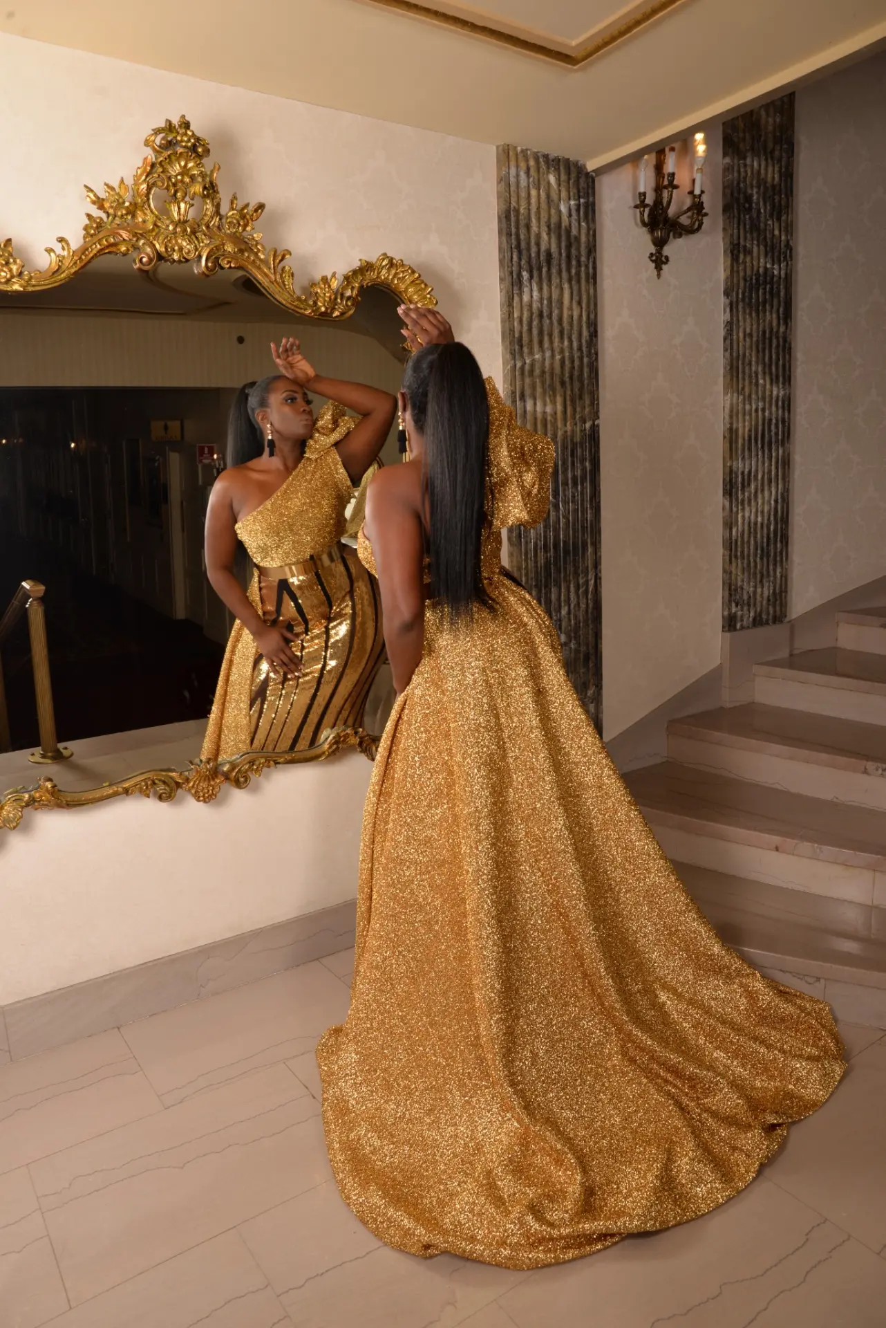 Gold one-shoulder glitter gown with a ruffled sleeve and sweeping train, beside a gilded mirror