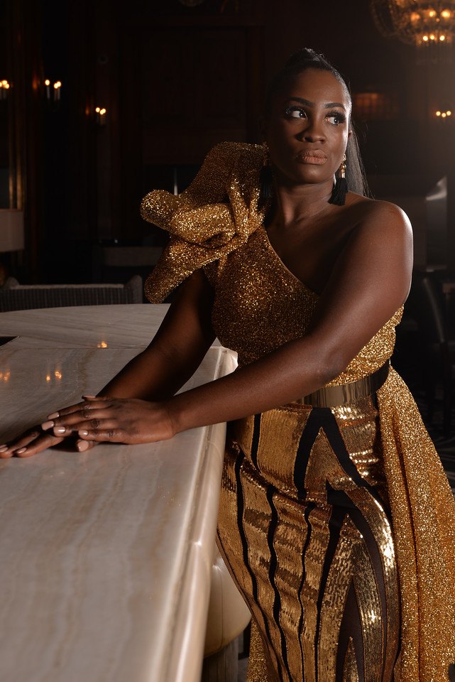 Metallic gold one-shoulder gown with a sculptural shoulder puff, a gold mirror belt and a glittering cape train, leaning on a marble bar counter