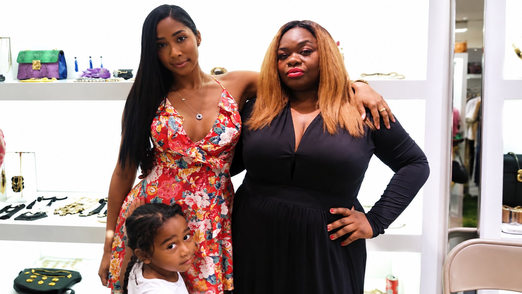 Therese Marie in a red floral halter dress with layered gold necklaces, posing with a guest in a black maxi dress at a boutique showcase of handbags and jewellery