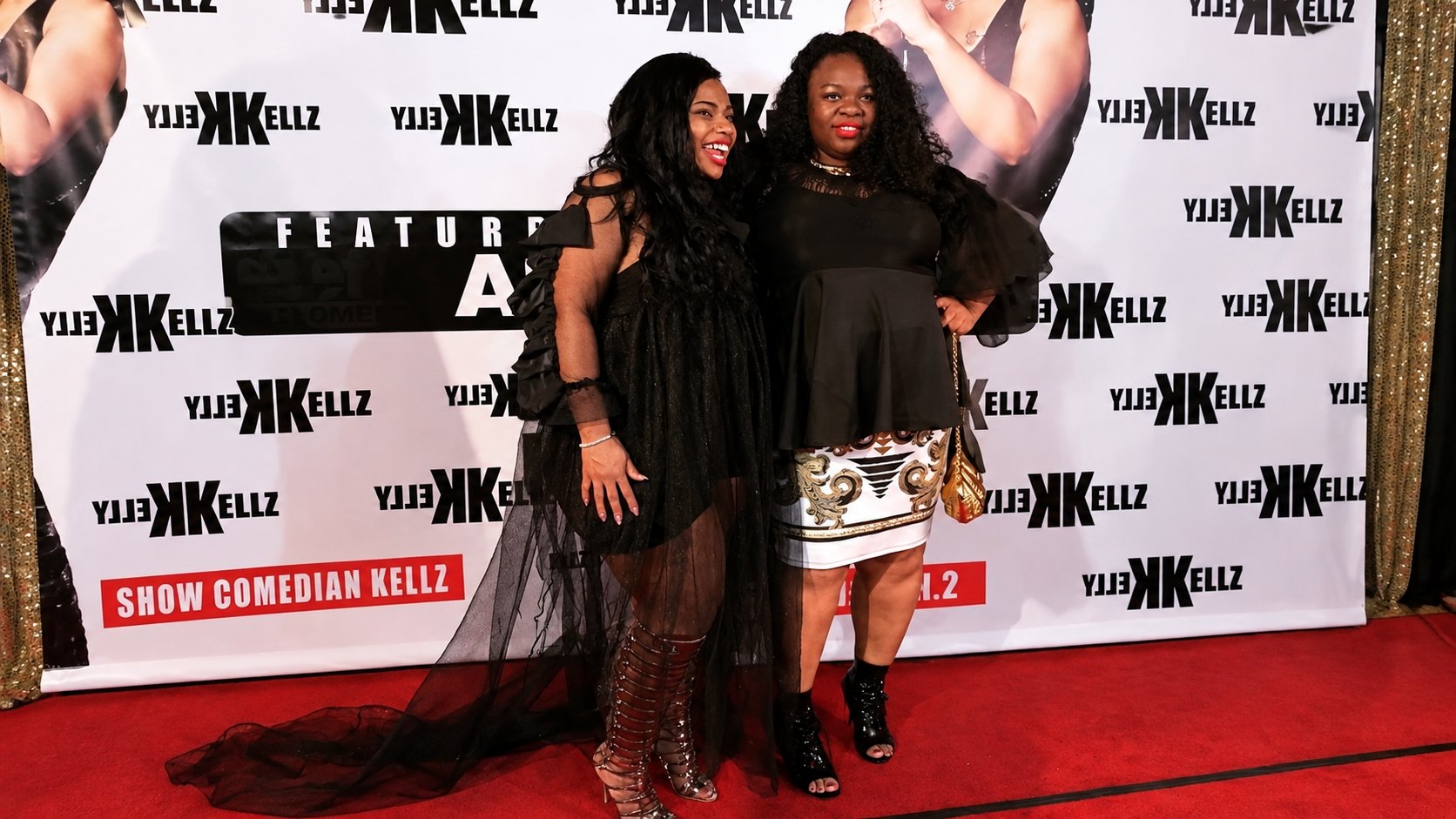 Therese Marie in a black mini dress with a sheer tulle train, posing with a guest in a white and gold patterned skirt on a red carpet photo wall
