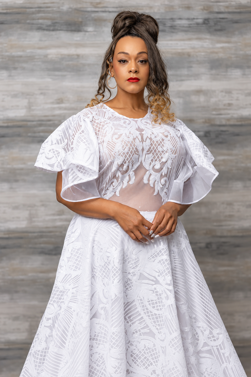 Model in a white gown with sheer mesh panels, floral lace embroidery and ruffled sleeves, posed against a grey weathered-wood backdrop