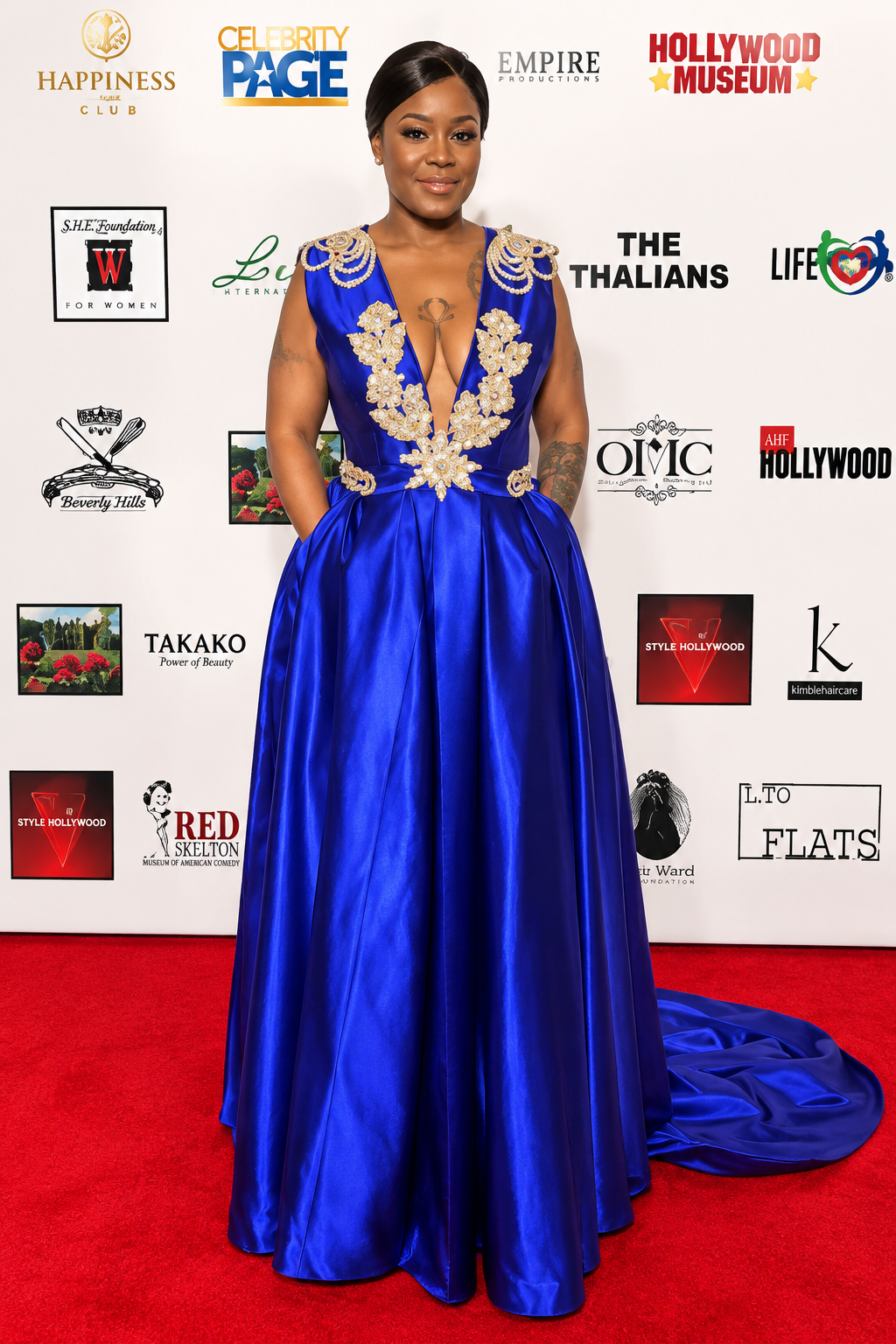 Sleeveless floor-length royal blue satin ballgown with a deep V neckline, gold and pearl beadwork on the bodice, pleated skirt with side pockets and a small train, on a red carpet