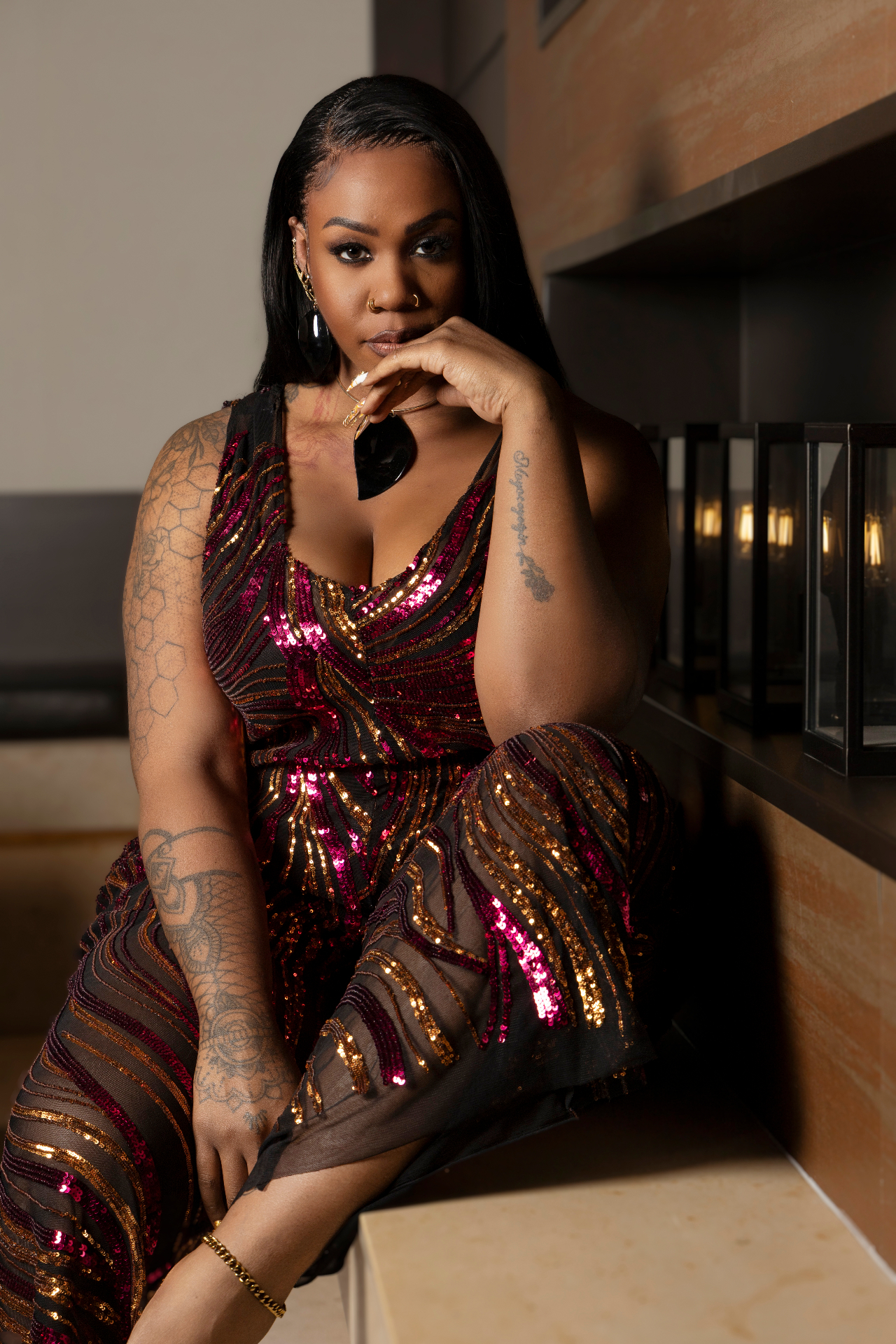 Close portrait in the magenta and bronze sequined mesh jumpsuit, seated in a warm alcove lined with glowing glass lanterns