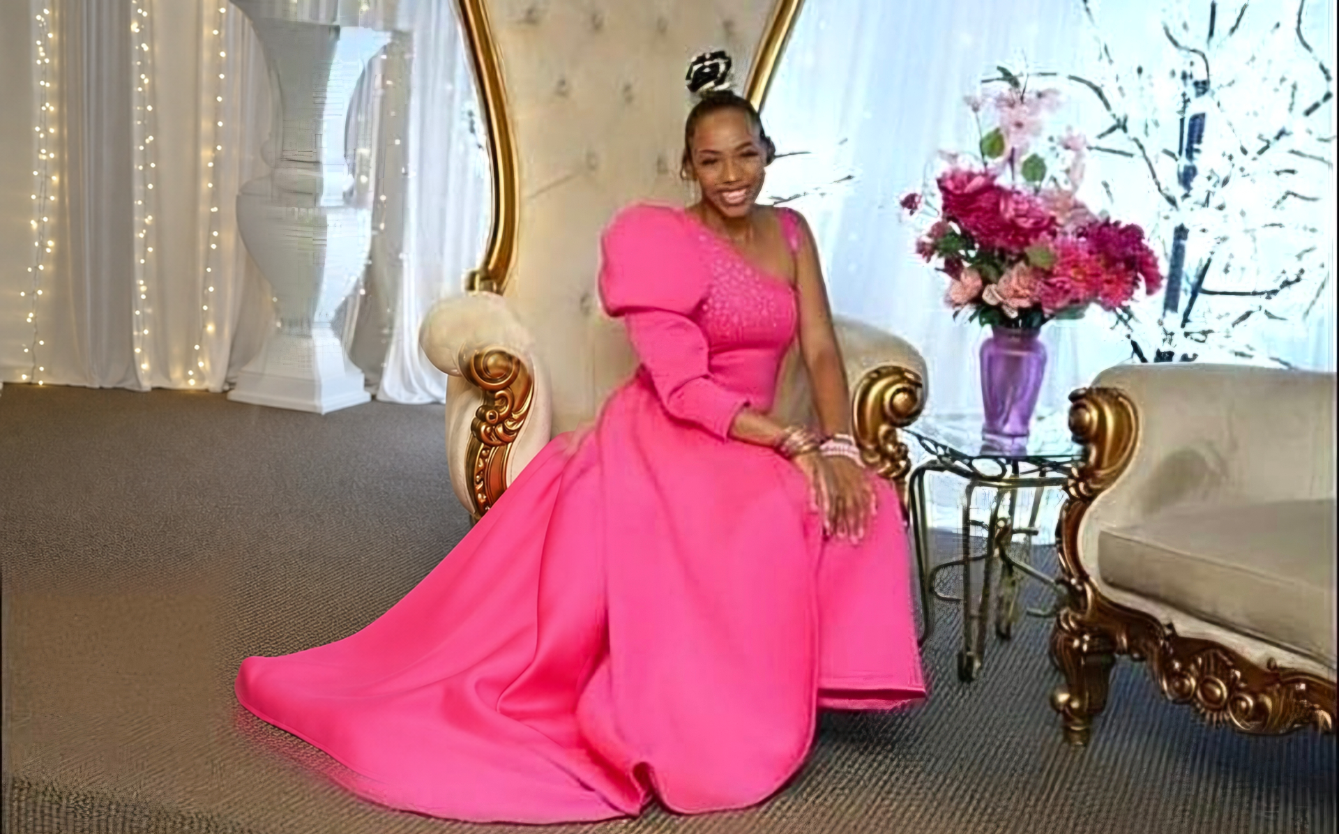 Model seated on a gold-trimmed throne chair in a hot pink one-shoulder gown with a beaded bodice, a puffed sleeve and a long flowing train