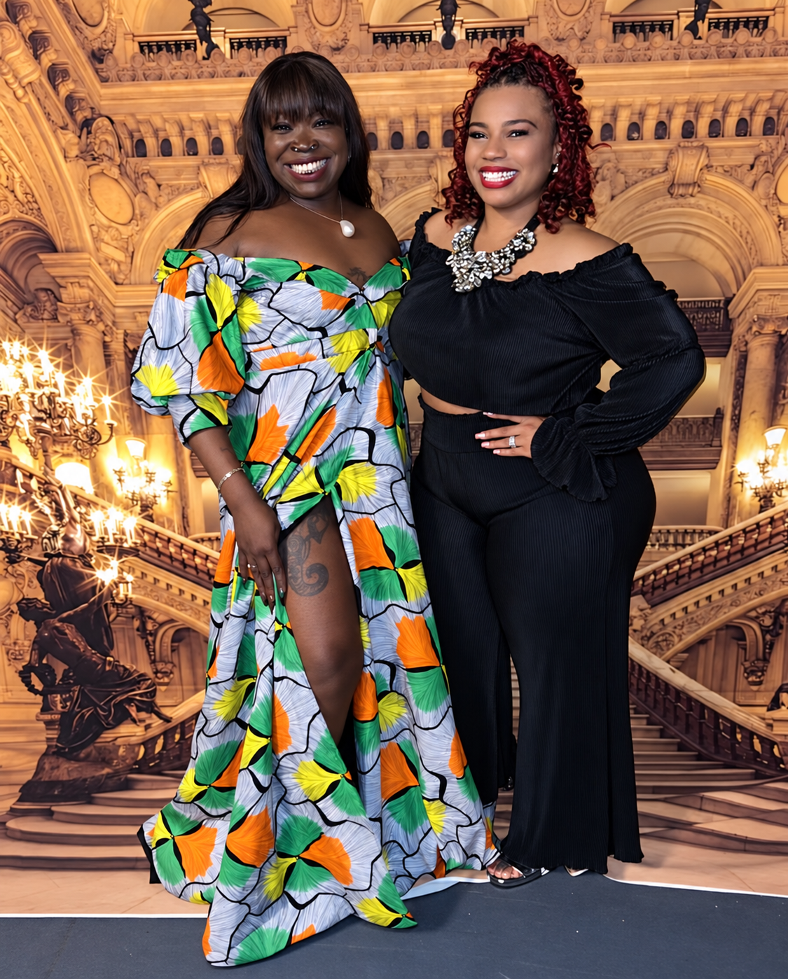 Two women together in front of a golden opera-house staircase backdrop — one in an off-shoulder grey, green, yellow and orange print gown with a high slit, the other in a black off-shoulder top and wide-leg trousers