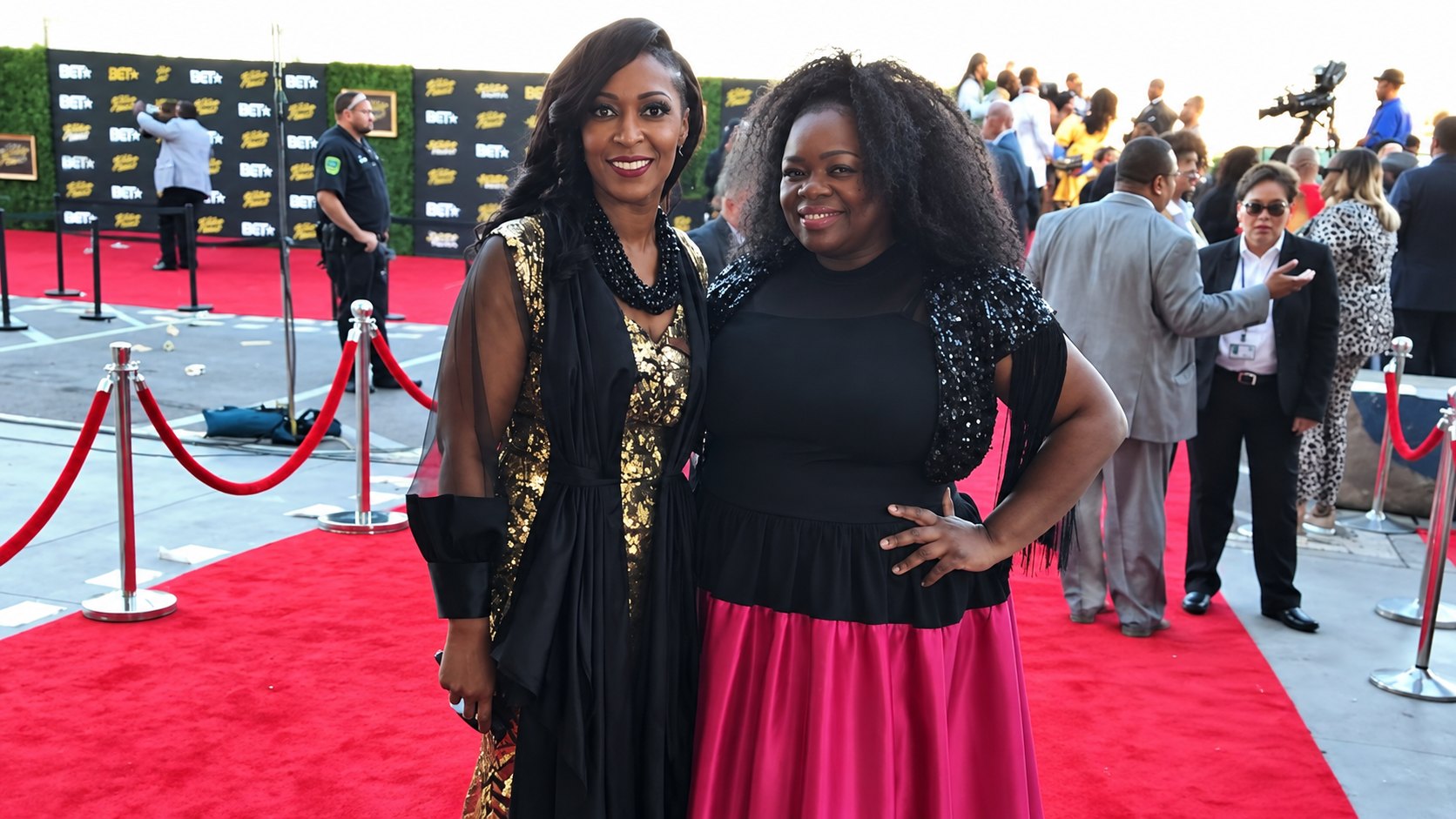 Therese Marie in a black and gold gown with a beaded statement necklace, posing with a guest in a magenta satin skirt on a red carpet