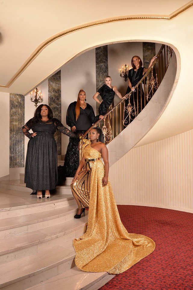 Five models on a curving marble staircase in a gold one-shoulder jumpsuit with a circular train, a black pleated gown, a black fringe gown, a black ruffled top with a gold striped skirt, and a black gown with a high slit