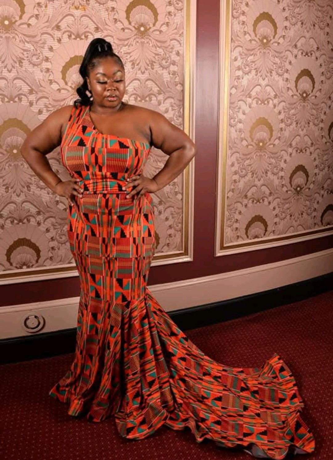 Orange and teal kente-print one-shoulder mermaid gown with a long sweeping train, against ornate gold-framed wall panels