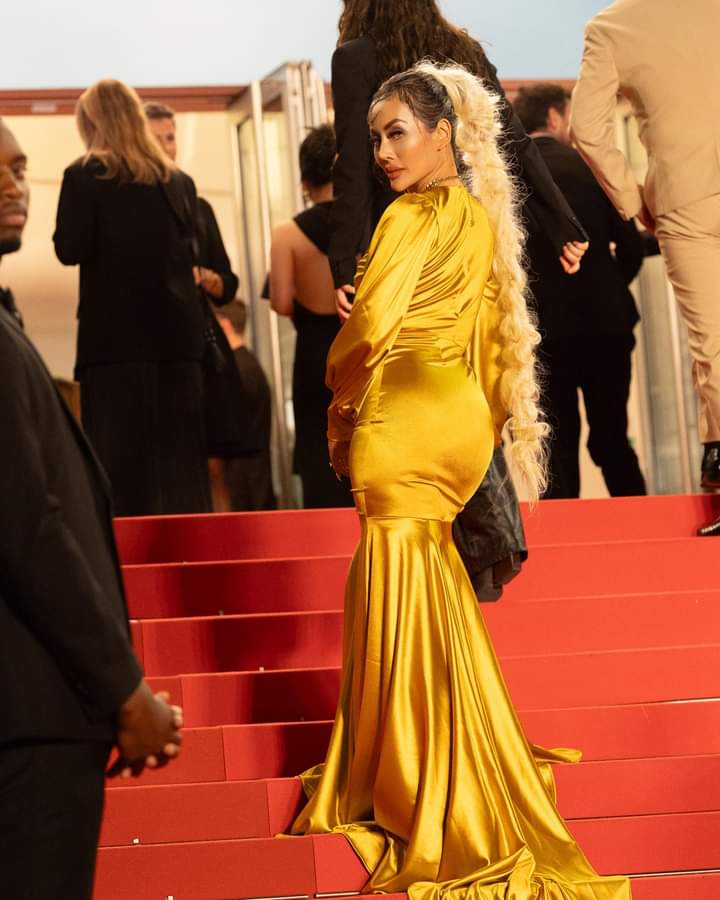 Gold satin long-sleeve mermaid gown with a floor-length train, on a red-carpeted staircase