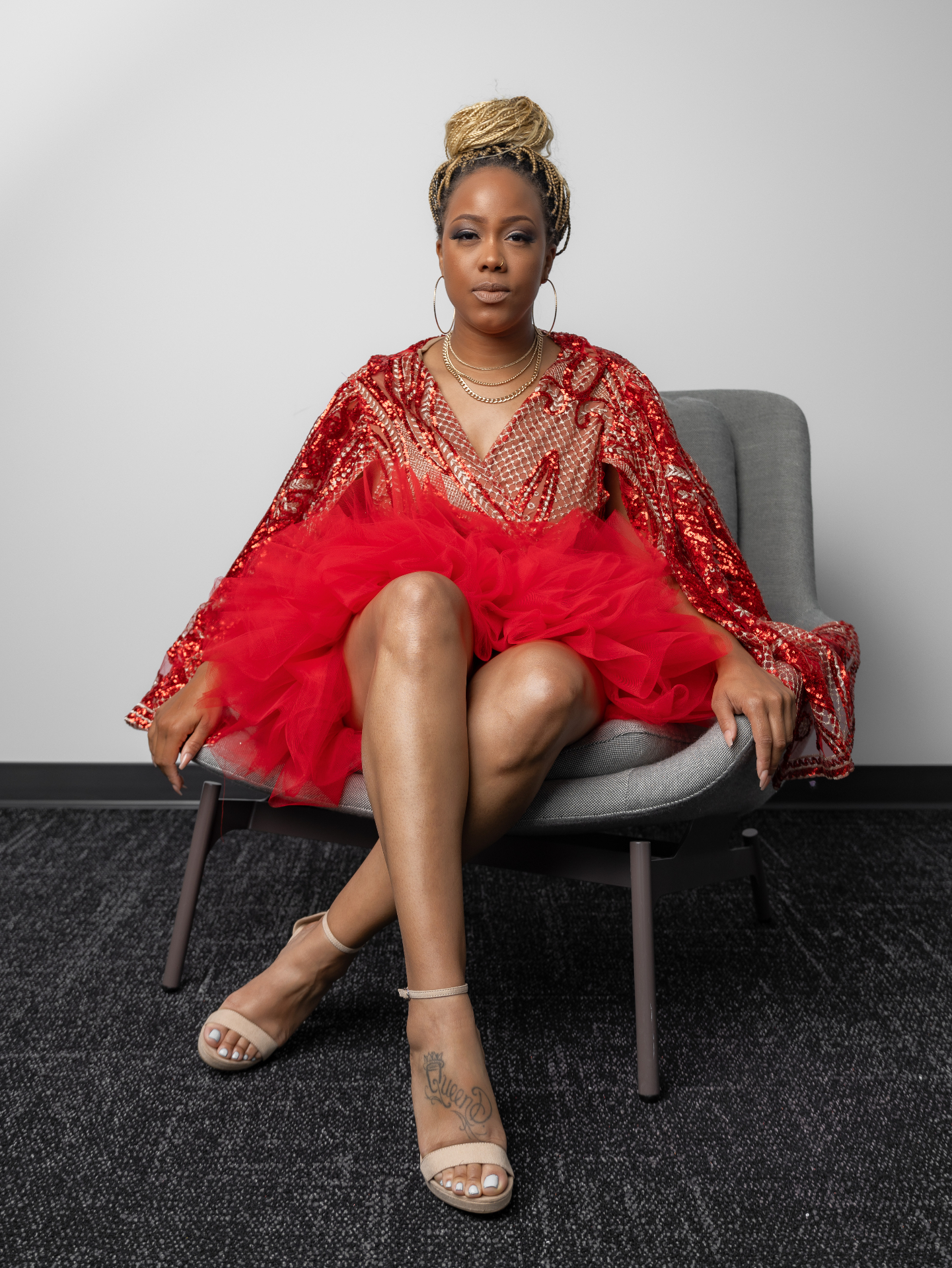 Red sequined cape overlay with geometric netting over an exaggerated red tulle puff skirt, seated in a grey armchair